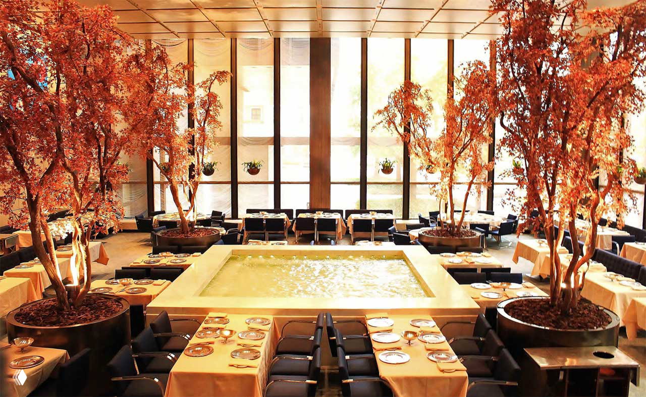 Роскошный зал ресторана Four Seasons с центральным водным бассейном и крупными декоративными растениями, столы с сервировкой и панорамные окна — атмосфера обеденного пространства.