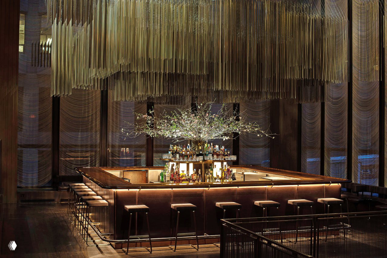Барная стойка в тёмном залитом мягким светом пространстве, над ней декоративная люстра-гирлянда и композиция со светящимися ветвями, стильный вечерний интерьер ресторана.