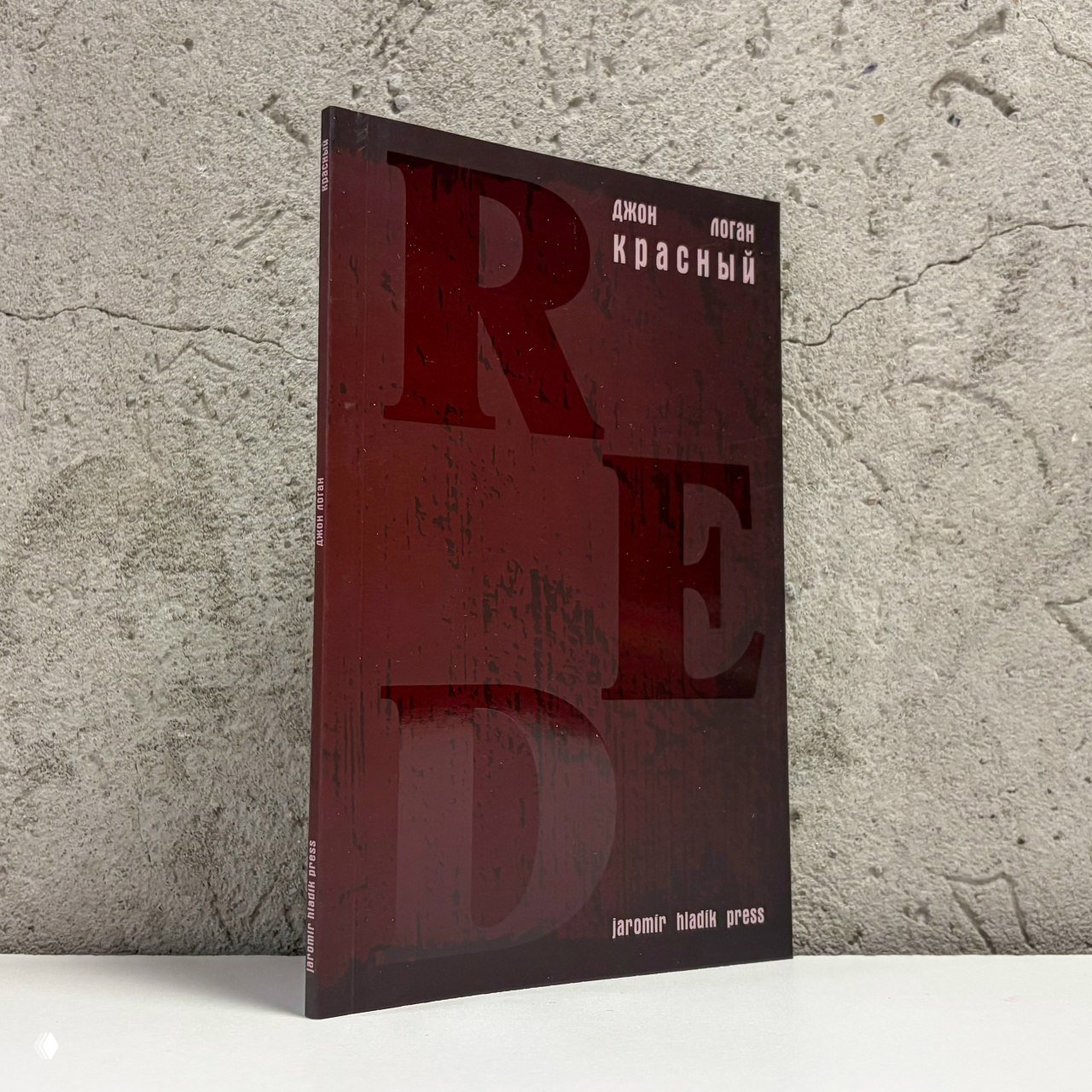 Фотография книжной обложки русского издания пьесы Джона Логана «Красный» от jaromir hladik press на фактурном фоне — издание в тёмно-красной гамме, фронтальный ракурс.