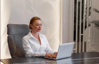 Женщина средних лет в белой рубашке сидит за столом с ноутбуком в светлом офисе; деловая поза и рабочая атмосфера, интерьер кабнета виден в кадре.