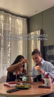 Таролог и клиент за столом во время гадания на суженого: карты, чашки и свечи в домашней обстановке с гирляндой на фоне.