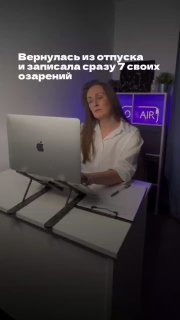 Женщина в рабочем кабинете за компьютером после отпуска: сидит за iMac, минималистичный офис и неоновая табличка «AIR», спокойная обстановка.