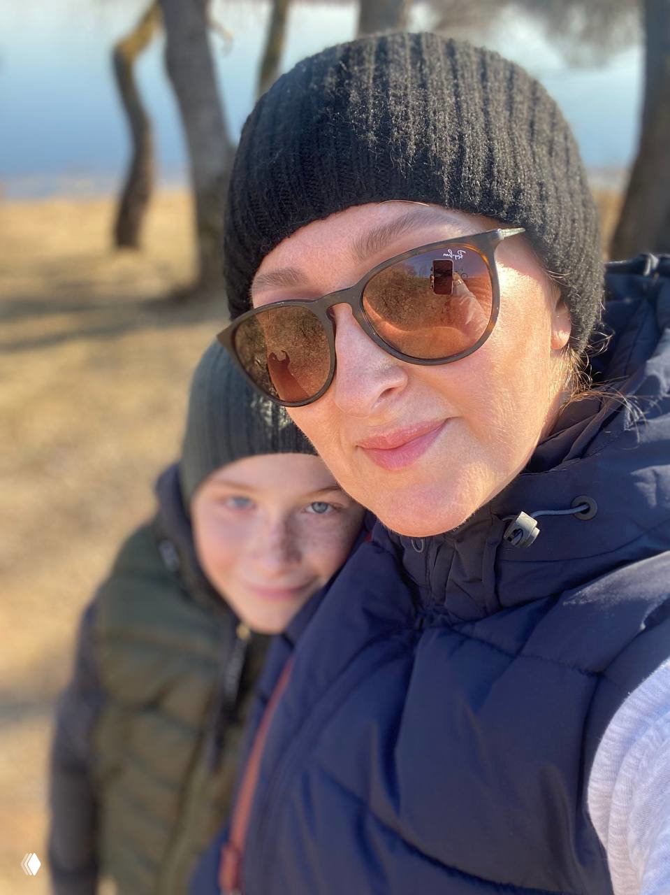 Крупный план женщины в одежде и ребёнка рядом на берегу озера; солнце, отражение в очках, тихая атмосфера.