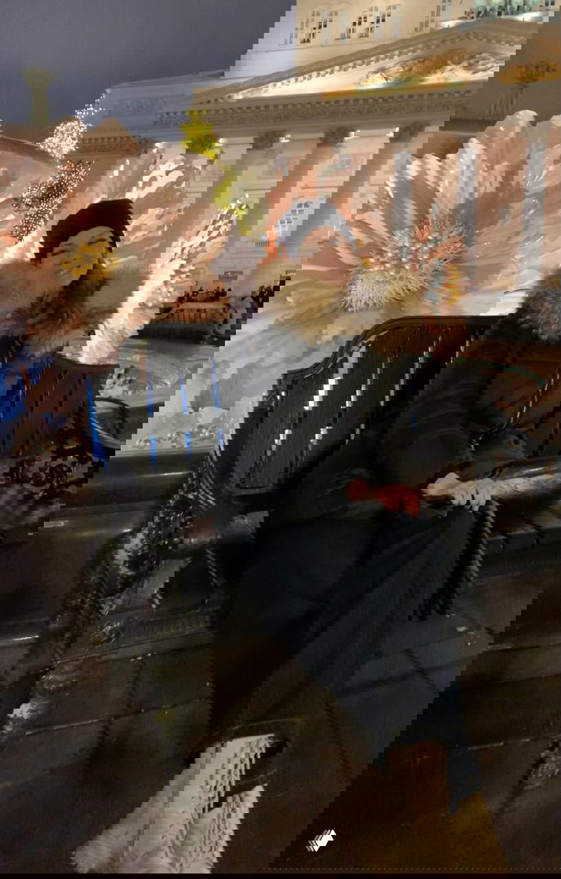 Автор на скамье в вечерней подсветке на фоне большой декоративной цветочной композиции и классической городской архитектуры в сумерках.