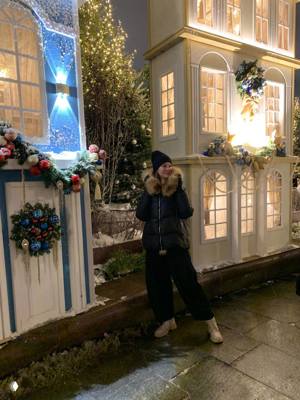 Женщина стоит возле белых и голубых рождественских домиков с гирляндами и хвойными композициями, на ногах видны зимние ботинки в снегу.