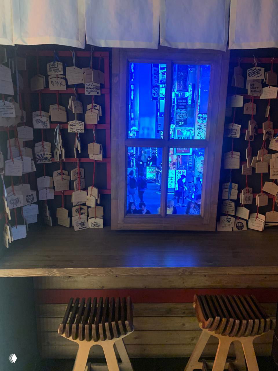 Деревянное окно с видом на неоновую улицу и вывески; вокруг висят дощечки пожеланий и стоят деревянные табуреты, атмосферный ночной кадр.