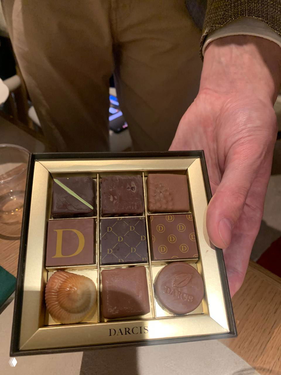 Открытая коробка с девятью шоколадными конфетами в золотой вкладке; коробку держат человеческие руки — вид разного ассортимента DARCIS.
