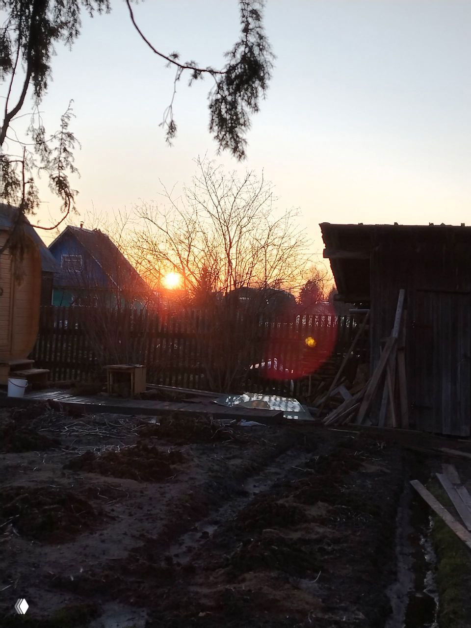 Вечерний закат над деревенским участком: солнце садится за домами, деревянные постройки, забор и силуэт деревьев на фоне неба.