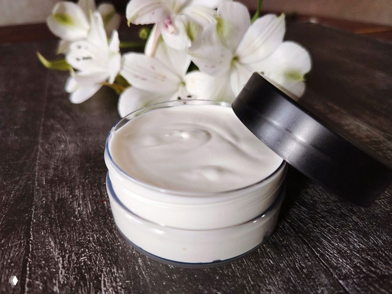 Открытая баночка с белым кремом на тёмном столе, позади белые цветы — крупный план натурального косметического бальзама или крем‑баттера.