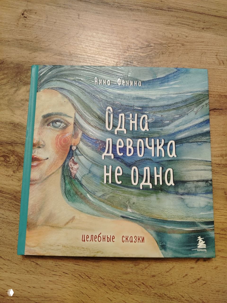 Обложка книги «Одна девочка не одна»: акварельный портрет женщины с развевающимися зелёно-голубыми волосами и крупным названием книги на фоне.