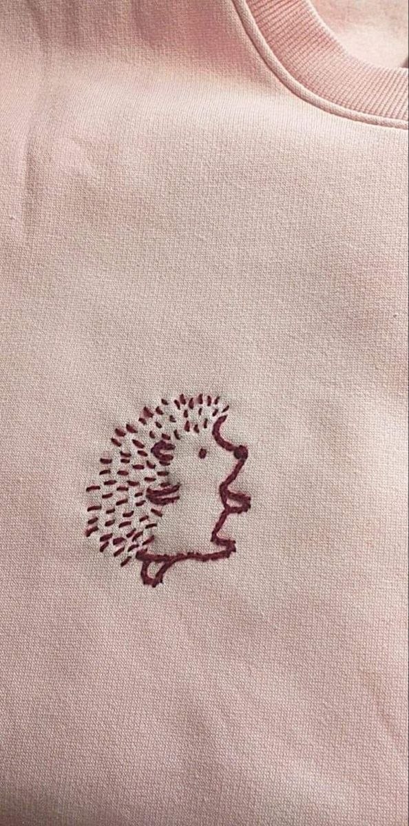 Крупный план розовой ткани со вышитым ёжиком тёмно‑бордовой нитью, пример аккуратной графической вышивки на одежде.