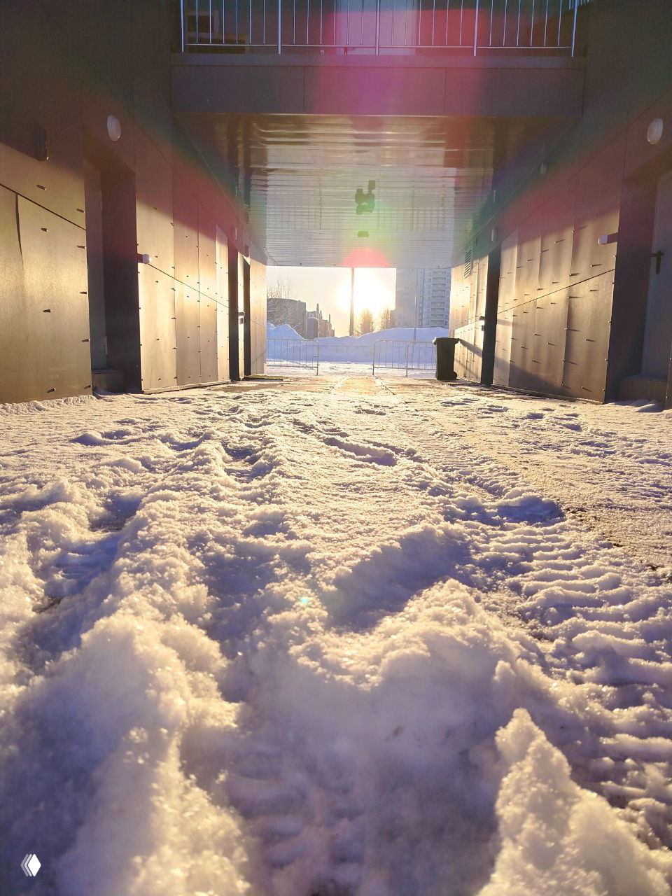 Низкий ракурс на снежную дорожку под аркой: следы на снегу, яркое низкое солнце на горизонте и радужные блики от солнечных бликов.