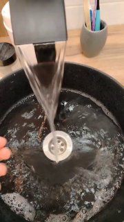 Рука держит твёрдый лавандовый шампунь под струёй воды над раковиной: видна струя, вращающаяся вода и мыльная пена в сером тазике, кадр демонстрирует процесс мытья шампунем.