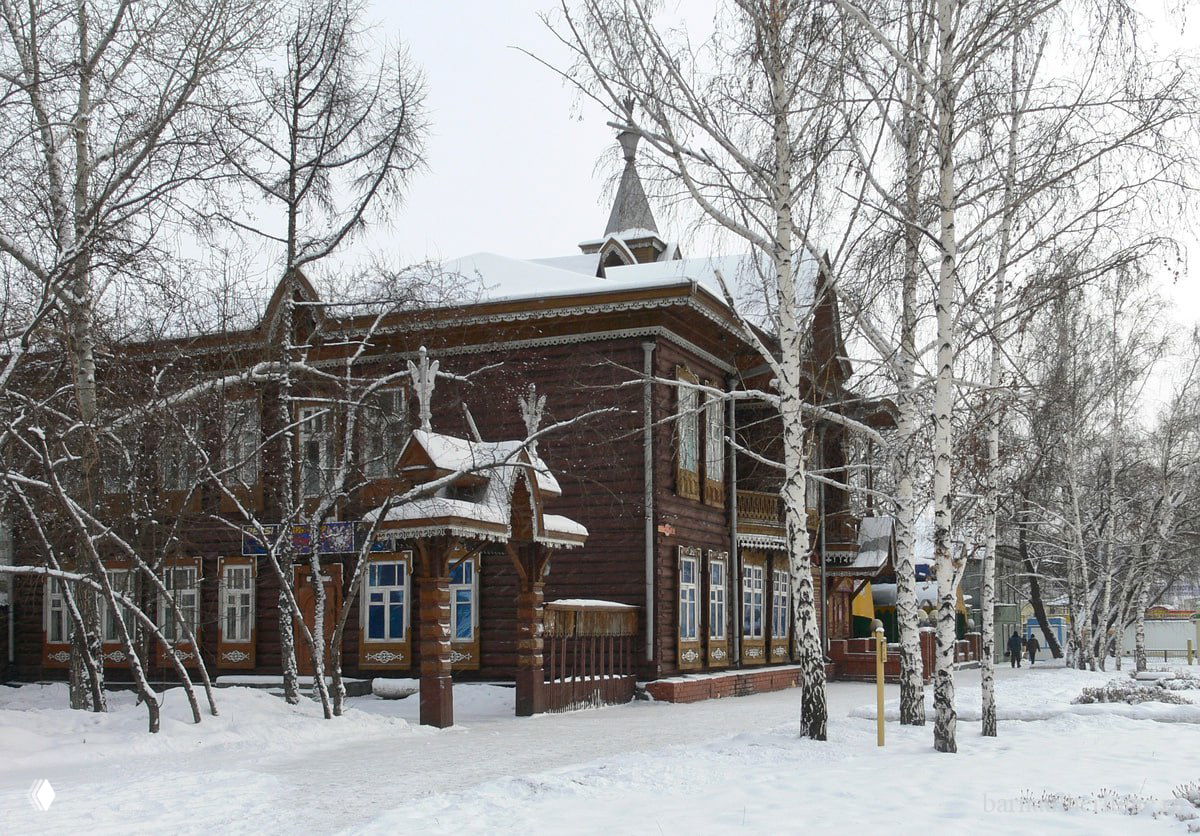 Фотография деревянной усадьбы Шадрина зимой: дом в снегу среди берёз, двухэтажный с резными наличниками и крыльцом, зимний пейзаж.