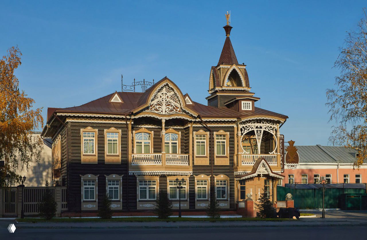 Полный вид деревянной усадьбы Шадрина в Барнауле: двухэтажный фасад с балконом, резными элементами и башенкой на фоне неба и осеннего света.