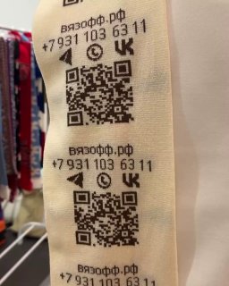 Крупный план вязаных бирок с QR‑кодом, номером телефона и иконками соцсетей на ткани — демонстрация читаемости кода на изделии.