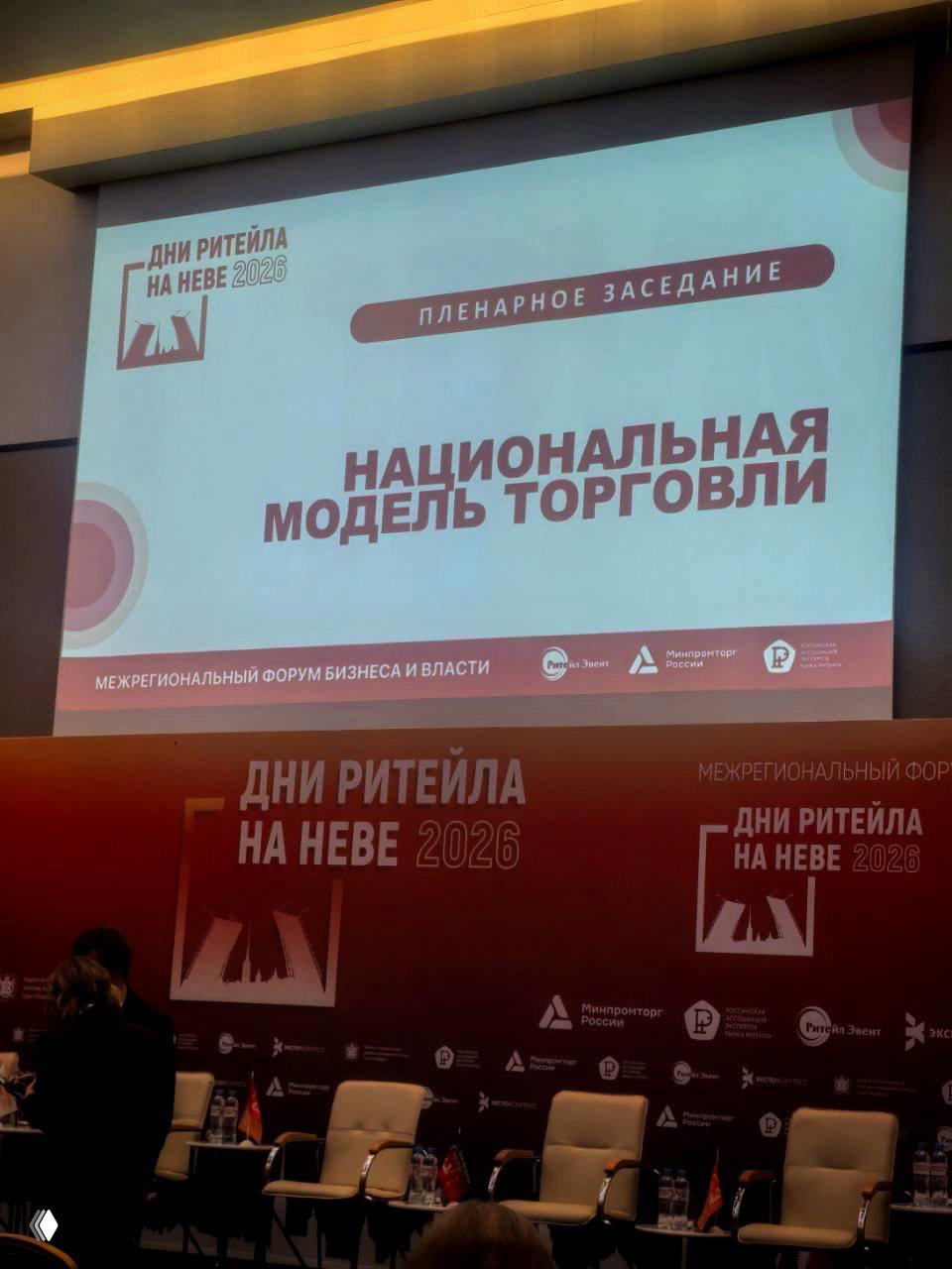 Кадр со сцены форума «Дни ритейла на Неве» — экран с заголовком пленарного заседания «Национальная модель торговли», вид на трибуну и подготовку к сессии.