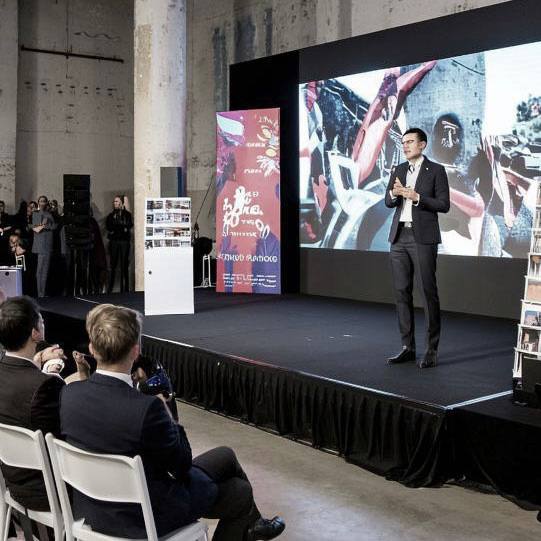 Сцена конференции: спикер с микрофоном выступает на фоне большого экрана, в зале видна аудитория и кресла — кадр с мероприятия/презентации.