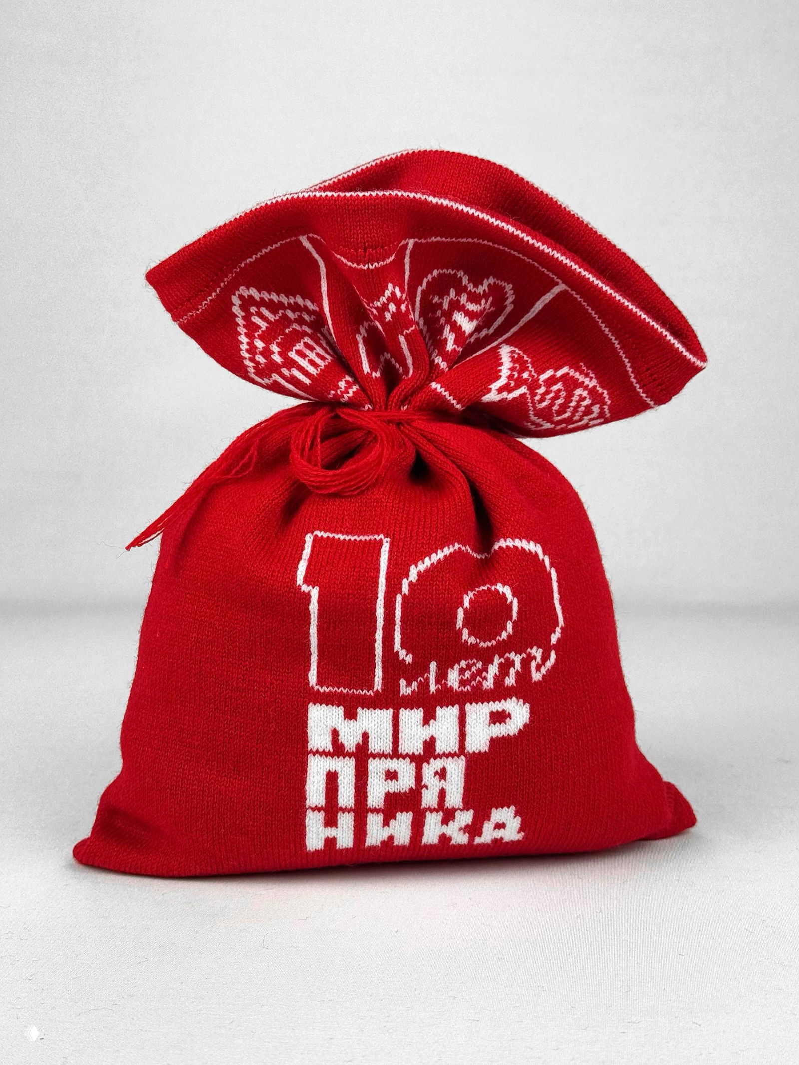 Красный вязаный мешочек с белым логотипом и надписью «Мир пряника», украшенный северным узором, сфотографирован на светлом нейтральном фоне.