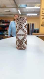 Вязаный чехол для бутылки стоит вертикально на столе в мастерской: виден орнамент, фактура ниток и полки с пряжей.