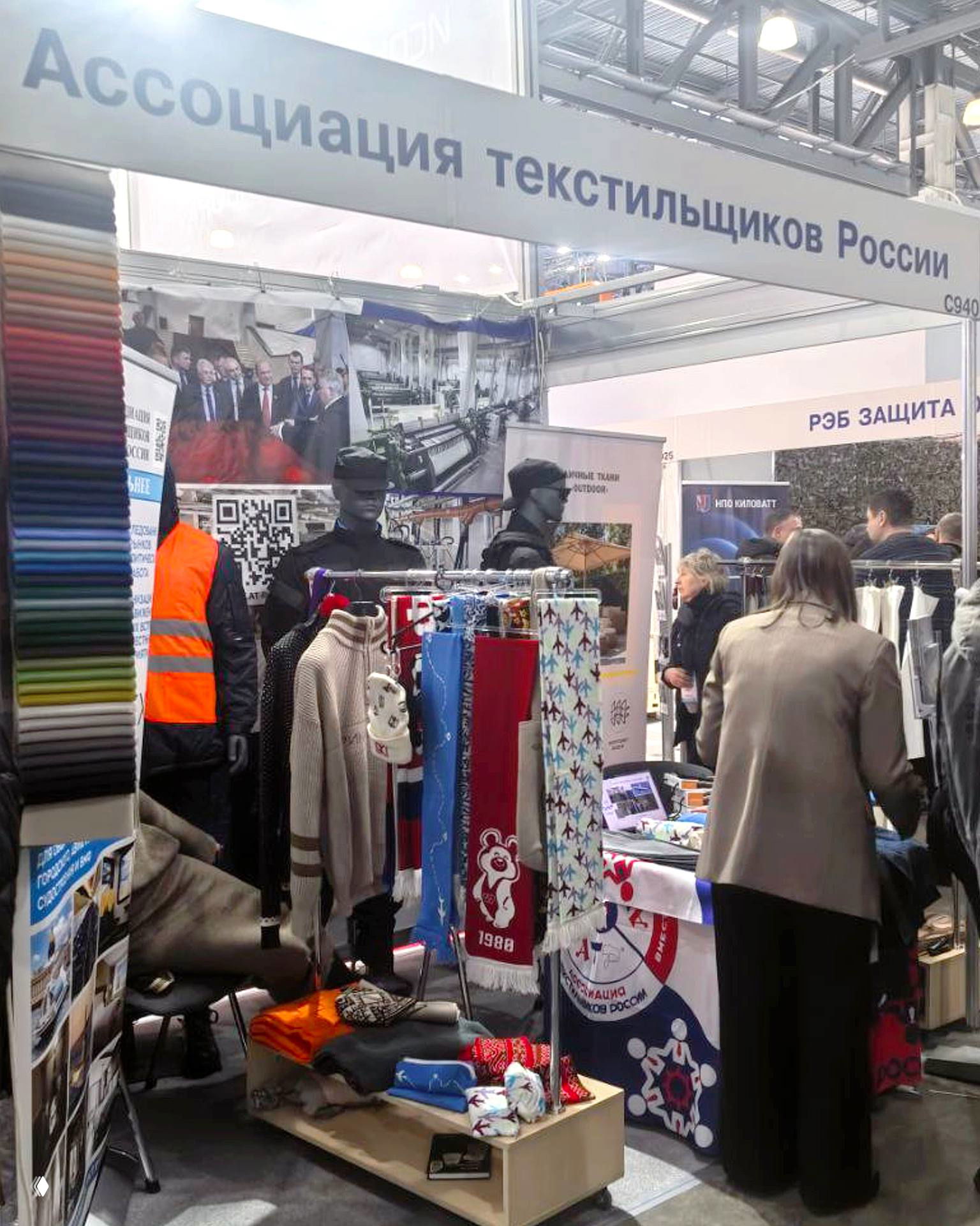 Стенд Ассоциации текстильщиков России с образцами трикотажа, шарфами и пледами; посетители осматривают изделия на НАИС 2026.