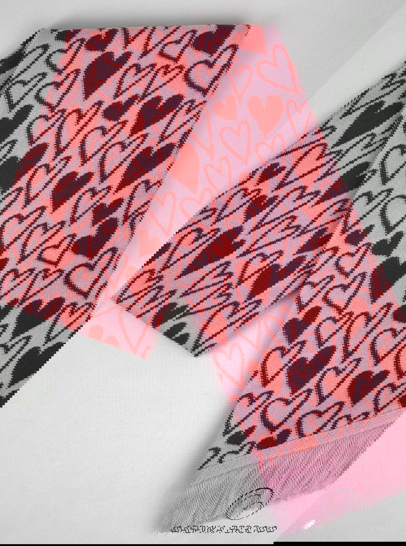 Розово-красный шарф с плотным узором сердечки, крупный план краев и бахромы, чёткая текстура трикотажа и фирменный логотип в уголке кадра.