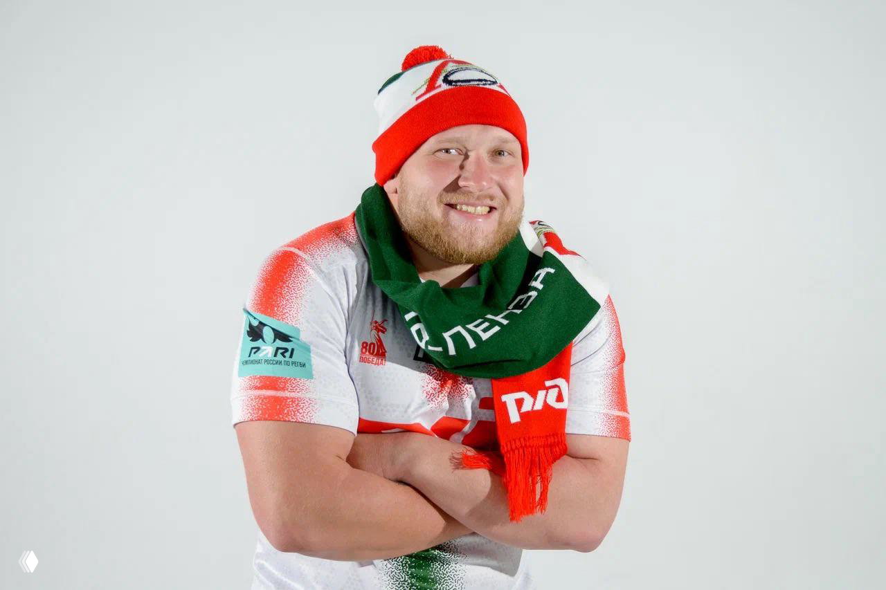 Игрок улыбается в красно‑белой шапке и зелёно‑красном шарфе с логотипом, демонстрируя связанный мерч команды в антураже белого фона.