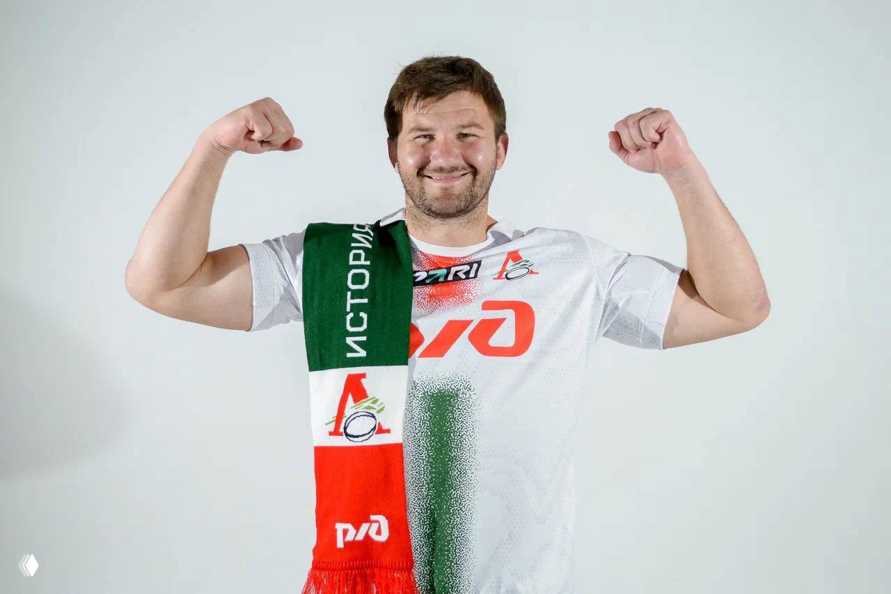 Игрок в белой футболке с логотипом и зелёно‑красным шарфом через плечо, позирует с поднятыми руками, презентация мерча в студии.