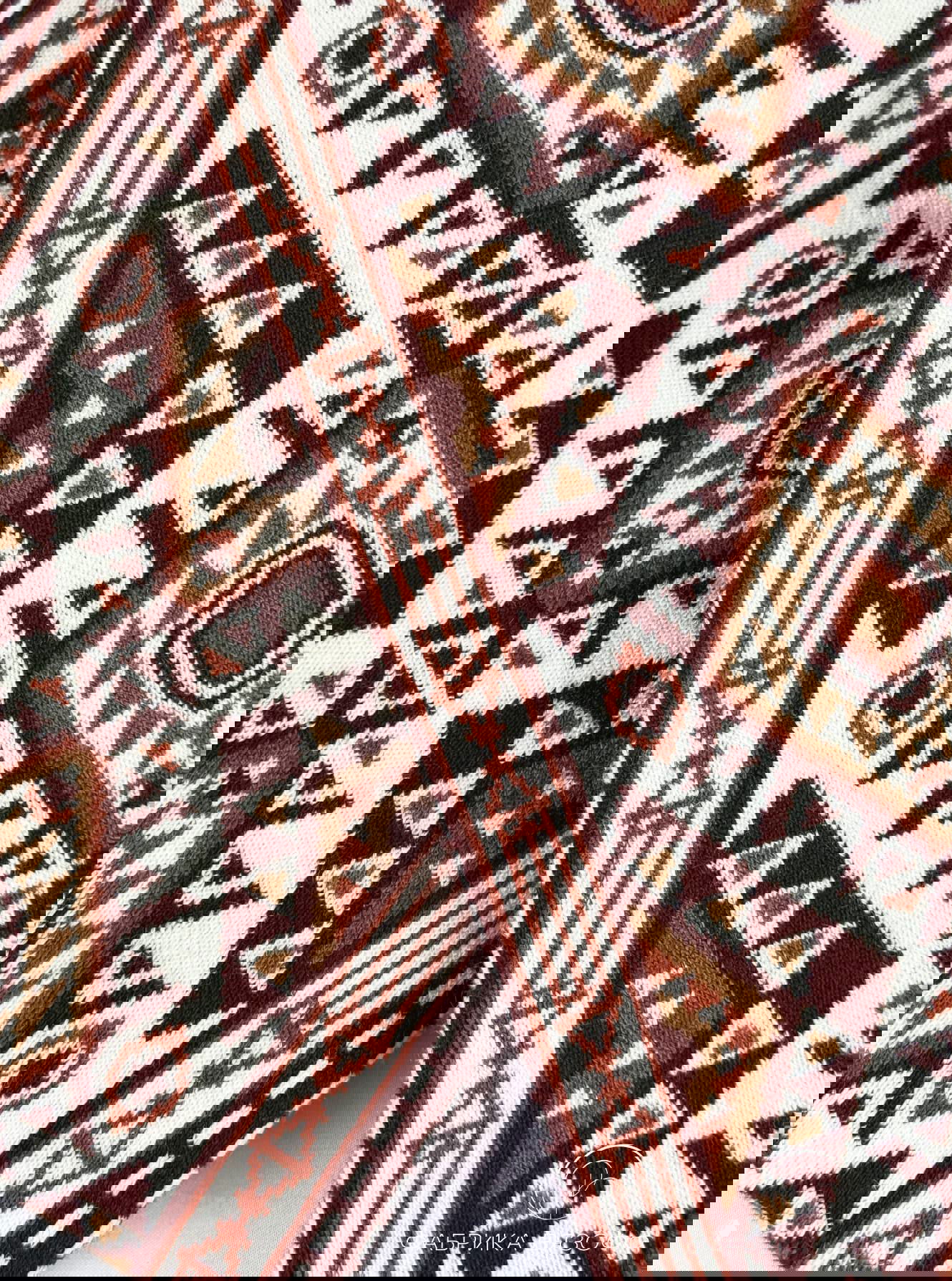 Крупный план детали шарфа с геометрическим орнаментом: белый фон, коричнево‑бордовые мотивы, текстура трикотажа и аккуратная кромка изделия.