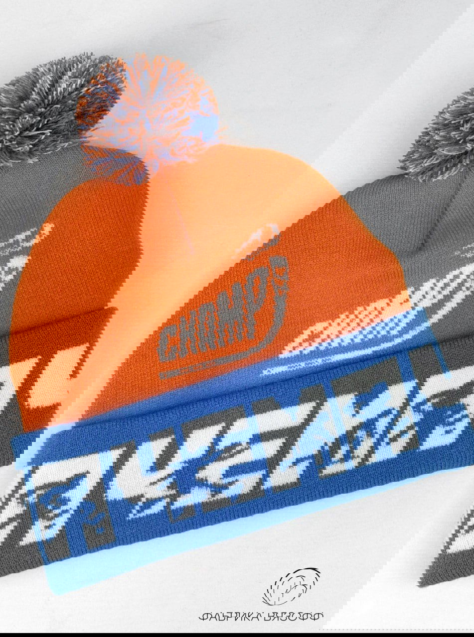 Крупный план шапки лёжа: ярко‑оранжевый верх, синий отворот с белыми элементами и помпон сверху, видно вывязанный логотип «Чемп».