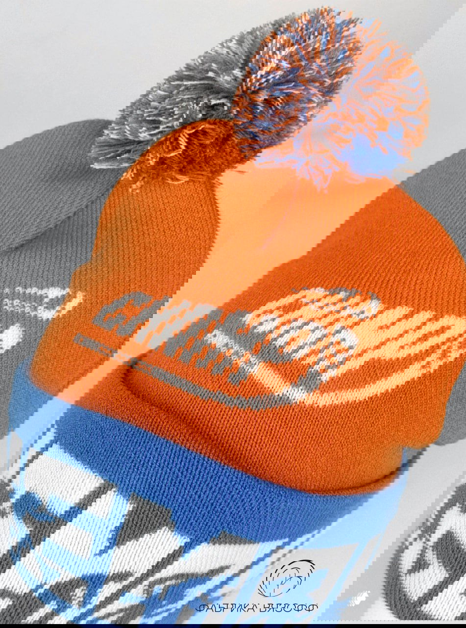 Детальный ракурс оранжевой шапки с белым логотипом «Чемп» и разноцветным помпоном, хорошо читается текстура вязки и плотность изделия.