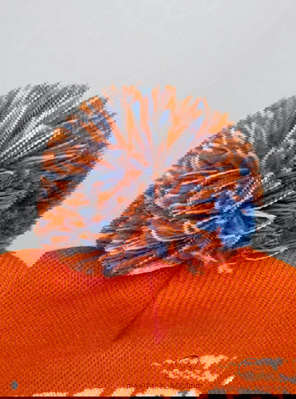 Крупный план помпона шапки: смешанные нити оранжевого, синего и белого цветов, фактура помпона и плотность пряжи хорошо видны.