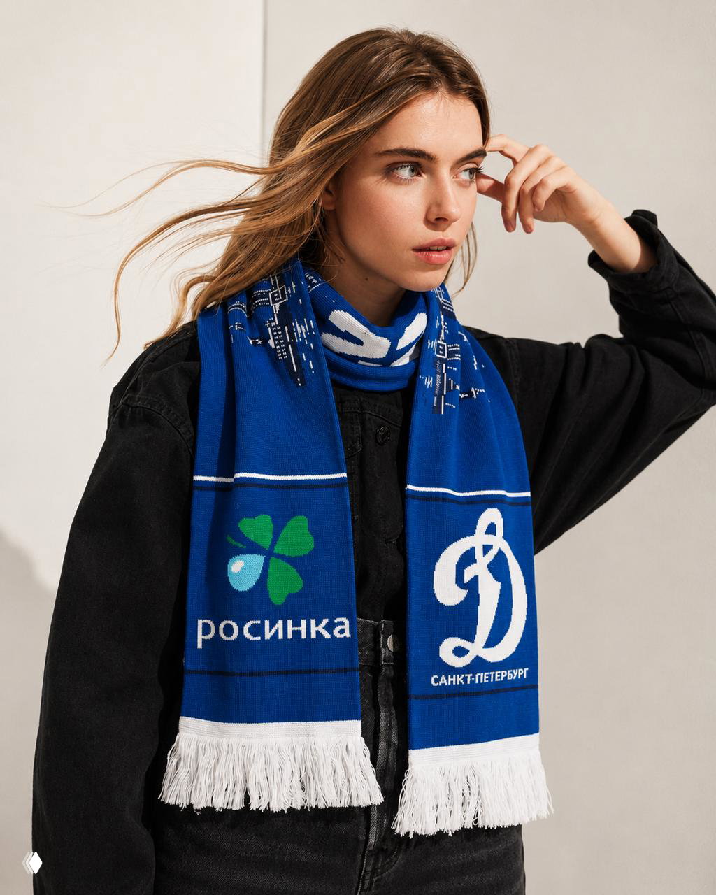 Женская модель в чёрной одежде позирует в синем вязаном шарфе с панорамой Санкт‑Петербурга и логотипами «Росинка» и «Динамо», шарф крупным планом.