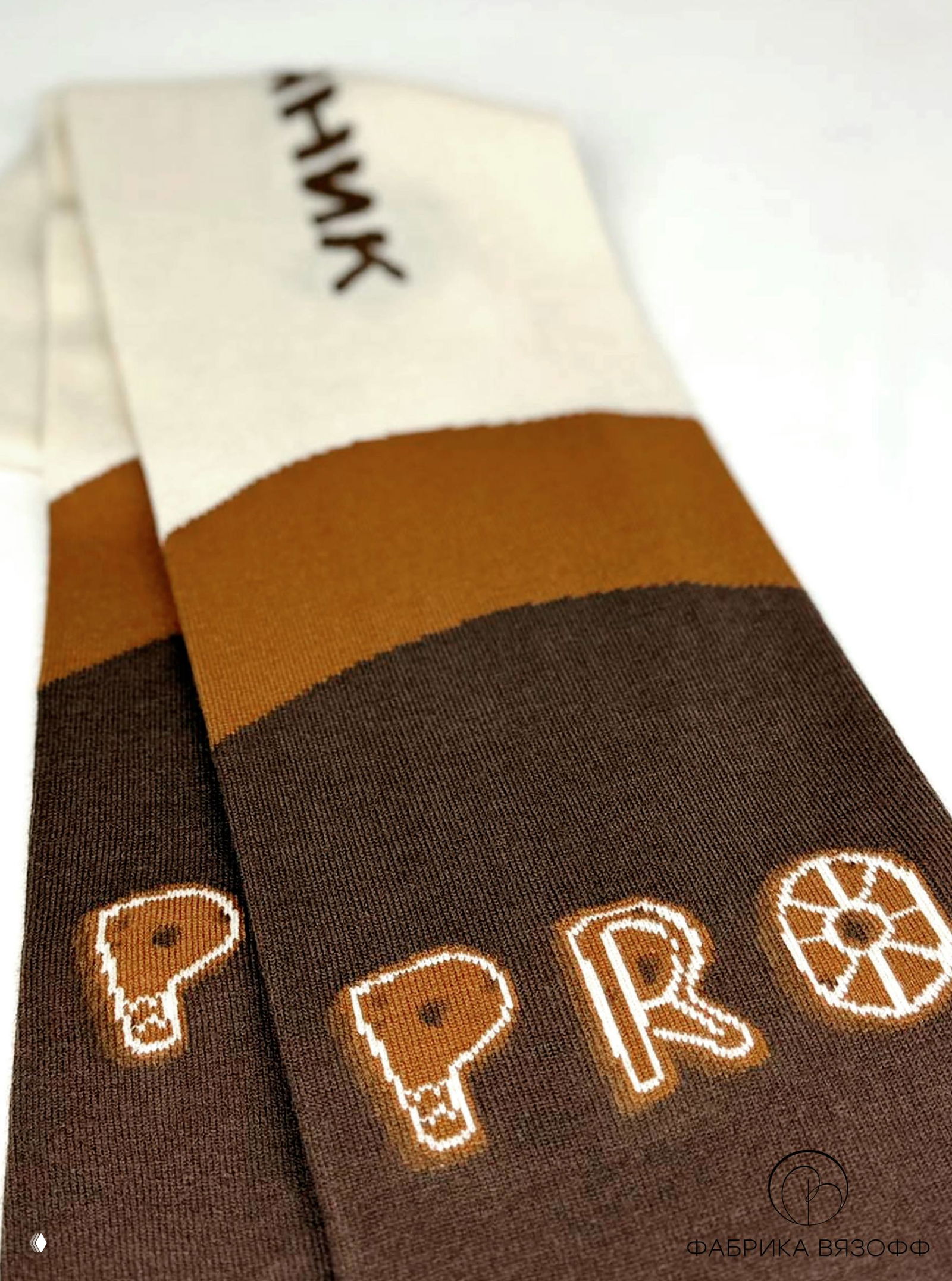 Детальный вид шарфа: перевёрнутый отрезок ткани с чётким рисунком букв, тёплые коричневые оттенки и гладкая фактура трикотажа на светлом фоне.