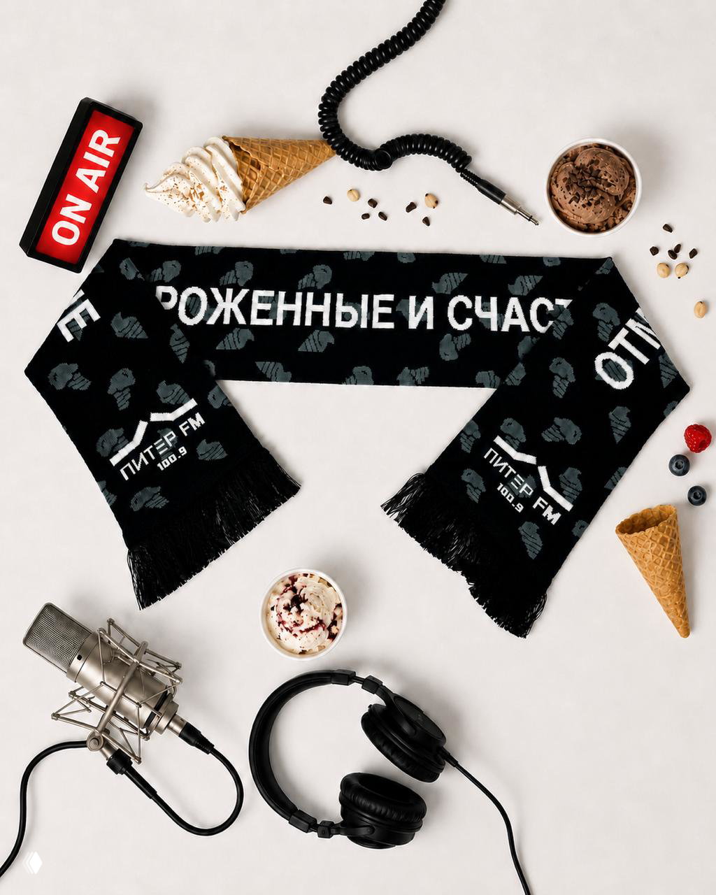 Чёрный вязаный шарф с широкой надписью «ОТМОРОЖЕННЫЕ И СЧАСТЛИВЫЕ», логотипом Питер FM и узором мороженого; раскладка с микрофоном, наушниками и рожками.