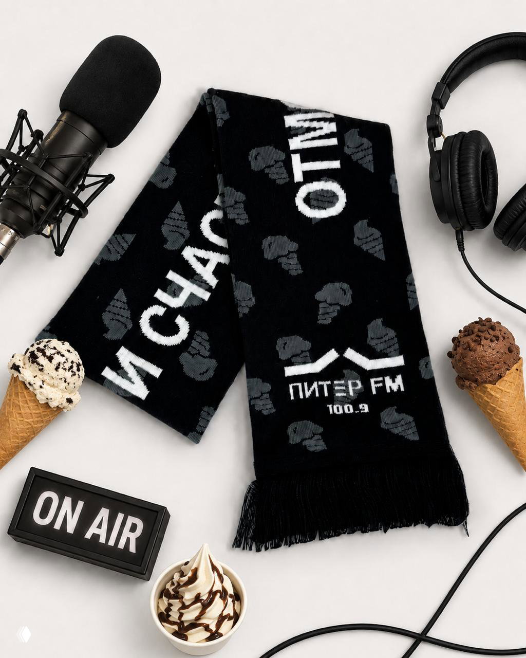 Крупный план чёрного шарфа с логотипом Питер FM и повторяющимся узором мороженого, рядом наушники и несколько порций мороженого в рожках для композиции.