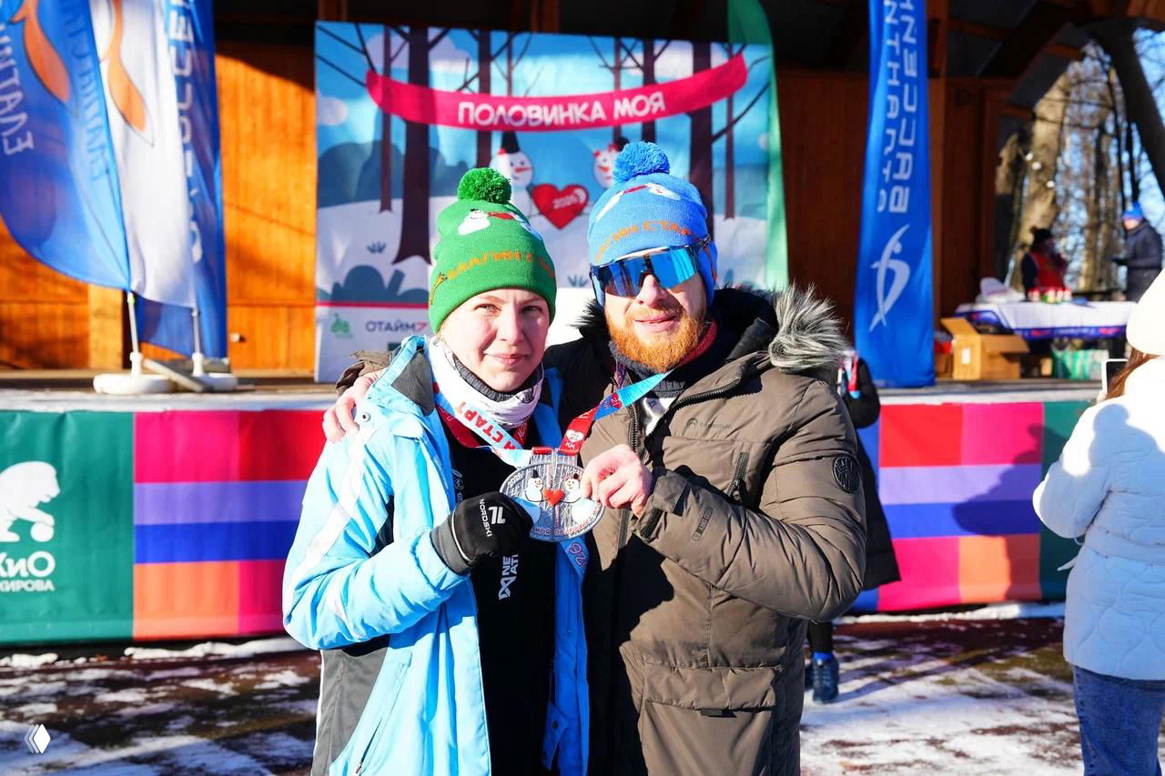 Участники забега на фоне сцены и баннера «Половинка моя», вязаные шапки с логотипом и снеговиками на головах, держат медаль после финиша.