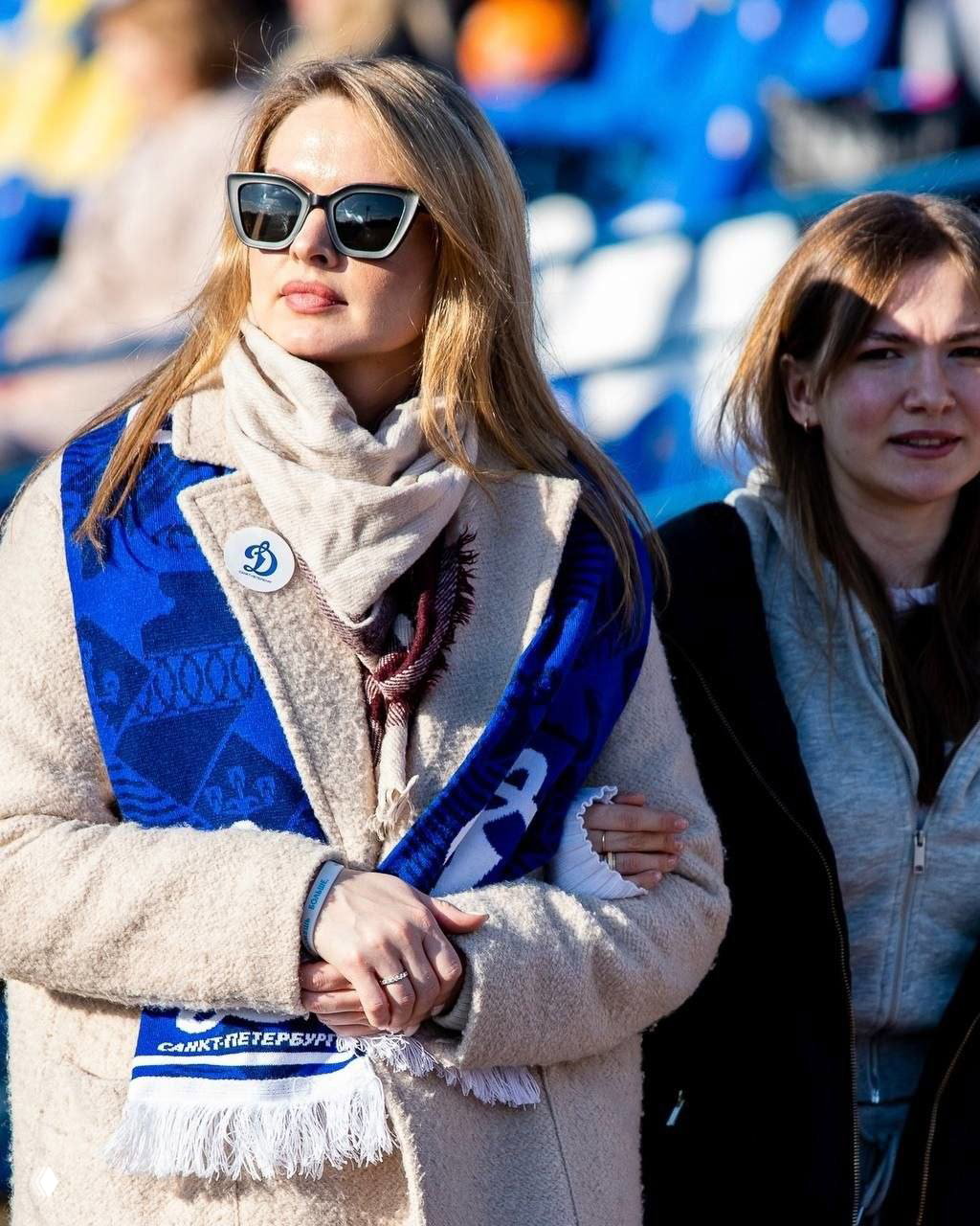 Женщина в солнцезащитных очках и бежевой шубе с ярко-синим шарфом «Динамо» на трибуне, размытый фон стадиона и другие болельщики.