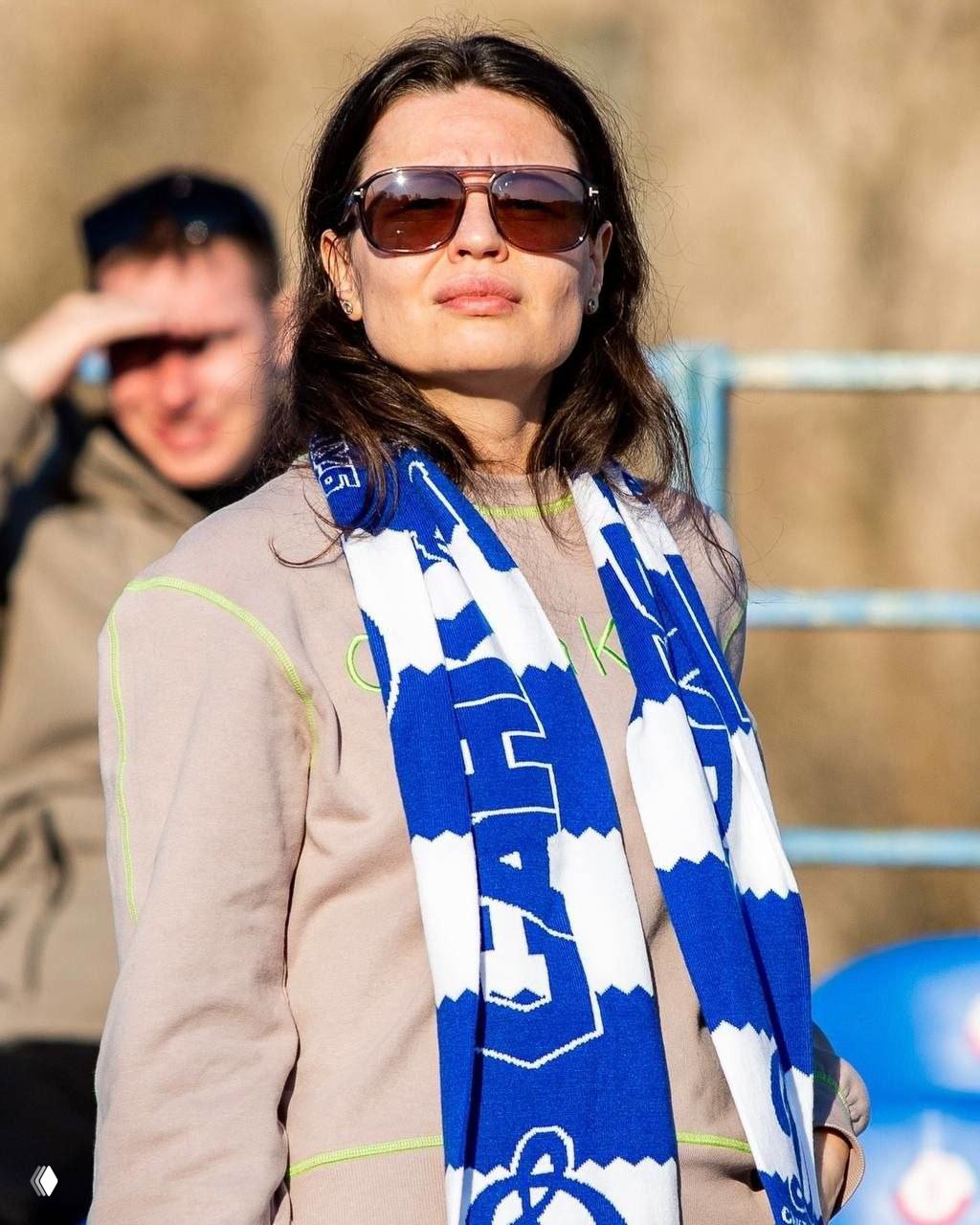 Портрет болельщицы в очках с длинными тёмными волосами, на шее — сине‑белый вязаный шарф с крупным узором и логотипом клуба, солнечный день.