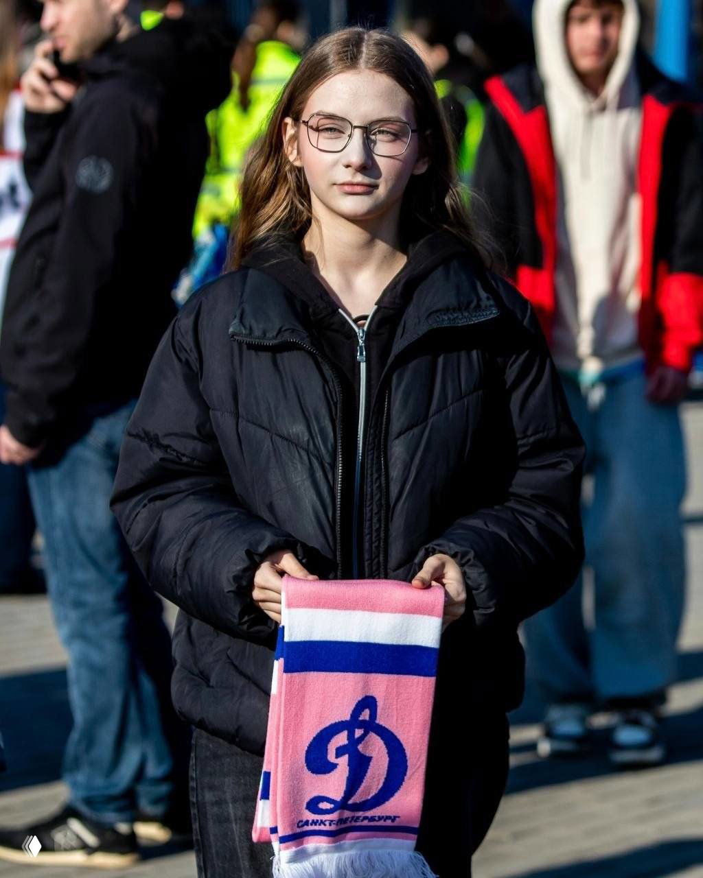 Подросток держит розово‑синий шарф с буквой «Д» и подписью «Санкт‑Петербург», на заднем плане толпа и элементы стадиона, уличная фотография.