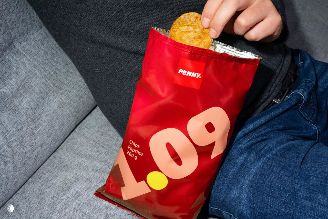 Красный пакет чипсов с большой розовой цифрой цены 0.09 на передней стороне; рука тянется за хлопьями из упаковки на диване.