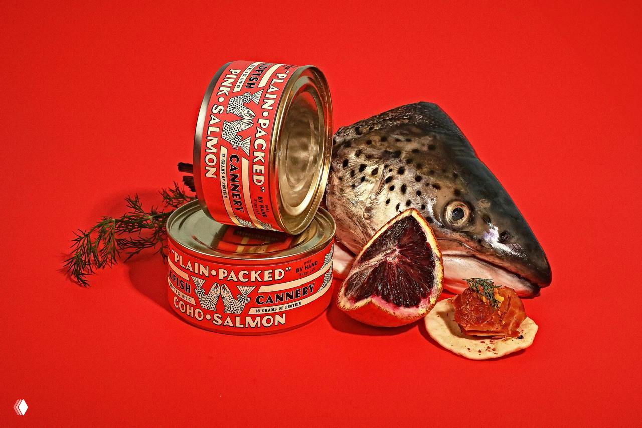Голова рыбы рядом с несколькими банками Wildfish и ломтиком цитруса на насыщенном красном фоне — художественная фуд-фотография с ретро-упаковкой.