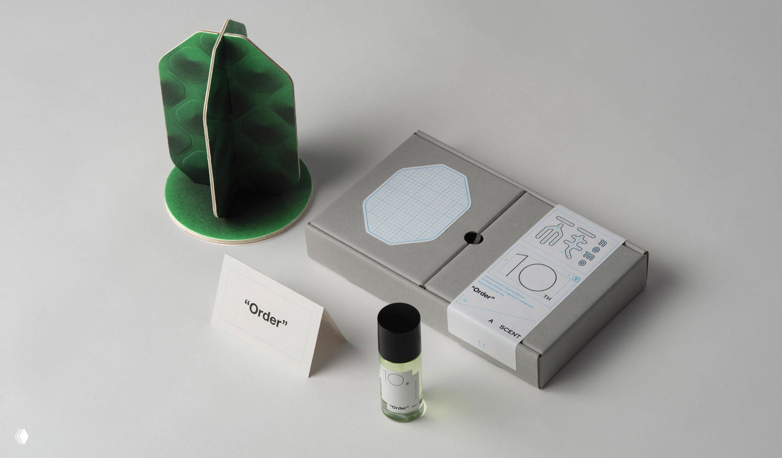 Съёмка на светлом фоне: раскрытая коробка, табличка с надписью «Order» и мини‑флакон духов рядом с текстурной зелёной конструкцией.
