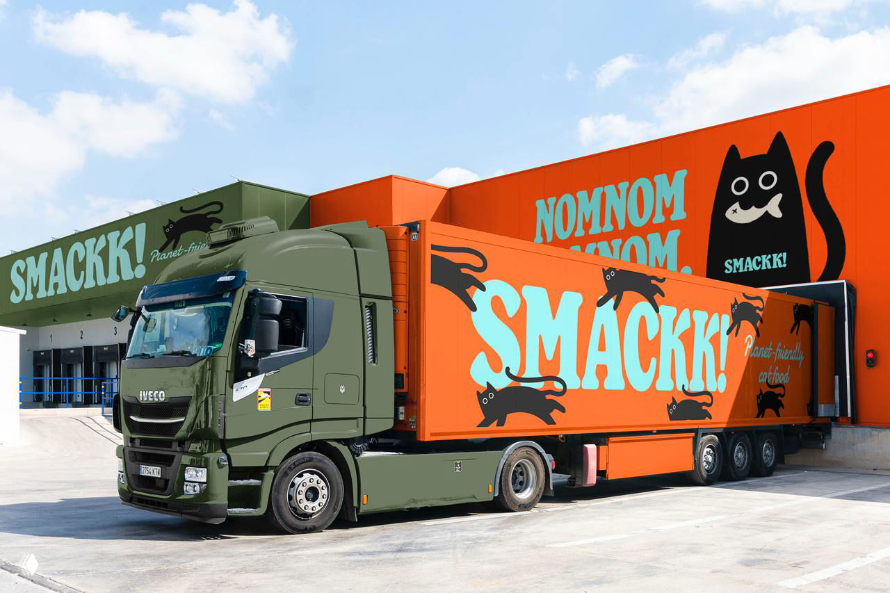 Грузовик и склад с брендингом Smackk: оранжевые и зелёные панели с большими буквами SMACKK и чёрными силуэтами котов на бортах.