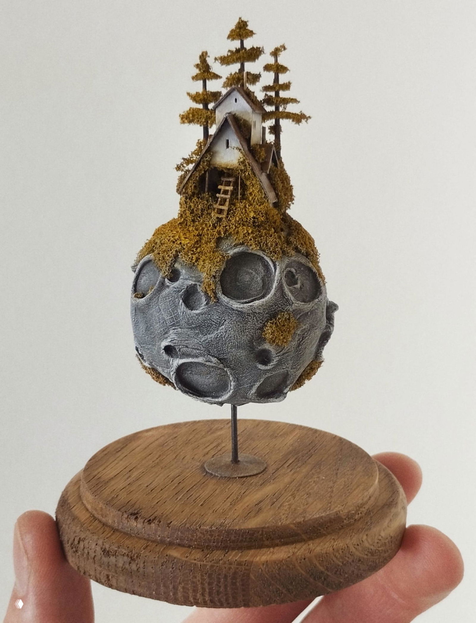 Миниатюрная планета со встроенным домиком «на вершине»: округлая, с кратерами и мхом, установленная на тонкой металлической стойке на деревянном основании.