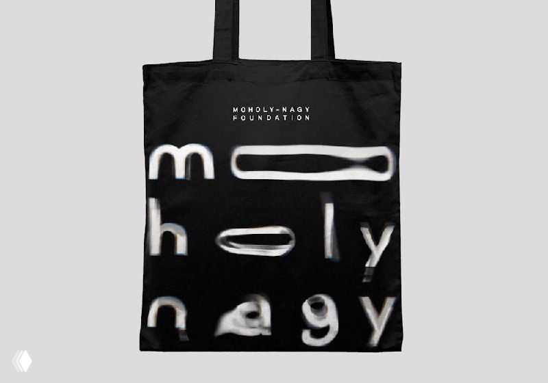 Чёрная сумка‑шоппер с фирменным нанесением Moholy‑Nagy: искажённая типографика на ткани, пример мерча и использования логотипа в носителях.
