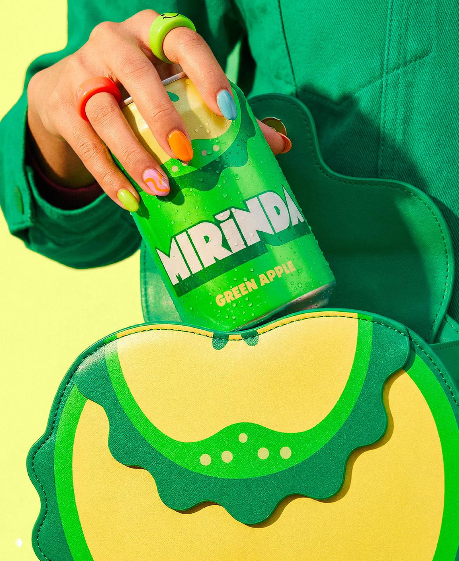 Рука с разноцветными кольцами держит банку Mirinda Green Apple на фоне зелёной сумки; рекламная съёмка подчёркивает свежую зелёную палитру и текстуру банки.