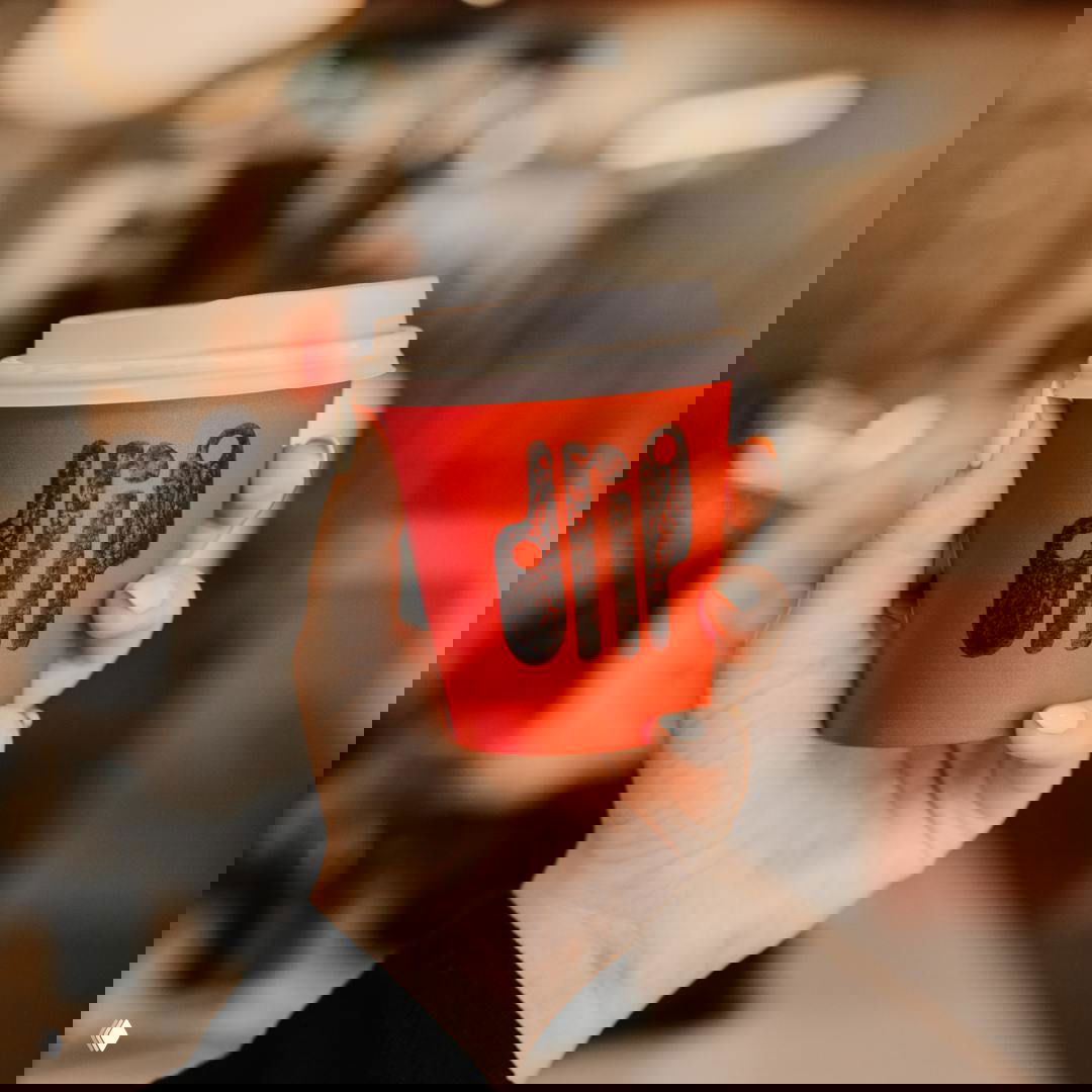 Рука держит красный бумажный стакан с отпечатанным логотипом Drip — выраженная текстура типографики и тёплый размытый интерьер на заднем плане.