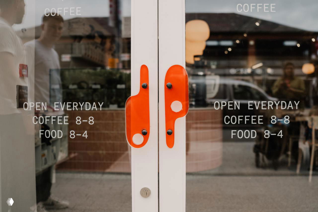 Двери кафе с оранжевыми ручками и надписями расписания работы: отражение улицы в стекле и частично видимый интерьер Drip за дверями.