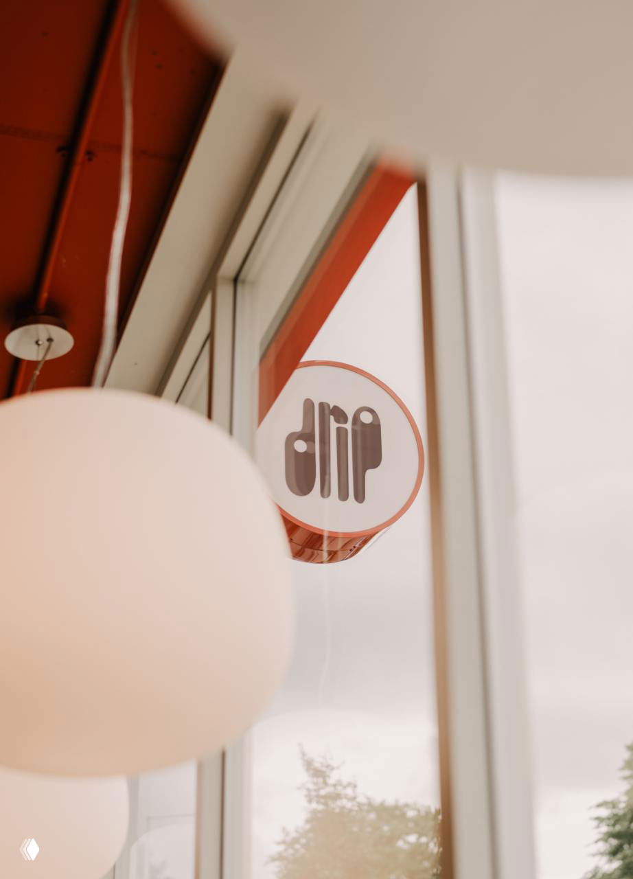Круглая вывеска Drip за стеклом, вид изнутри через витрину с крупными светильниками — ракурс подчёркивает фирменный знак и атмосферу кафе.